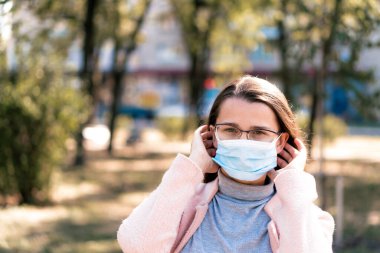 Bakım, bebekler, ilkbahar, koronavirüs ve karantina konsepti - Genç, uzun saçlı, Avrupa Kafkasyalı kadın, öğle vakti güneş ışığında mavi tıbbi koruyucu maske takıldı.