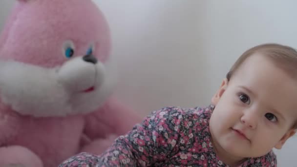 Αυθεντικό close up κοριτσάκι 1 έτους σε πολύχρωμο φόρεμα κάθεται και παίζει με ροζ αρκουδάκι σε παιδικό δωμάτιο σε λευκό φόντο σε εσωτερικούς χώρους. πατρότητα, παιδική ηλικία, έννοια της ζωής — Αρχείο Βίντεο
