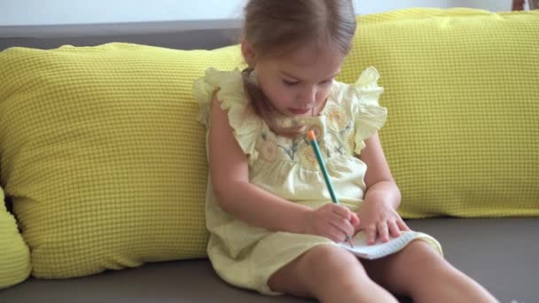 Portrait Petite fille drôle en robe jaune souriante enfant écrit avec un crayon dans un cahier assis sur le canapé à la maison. mignonne enfant femelle avec joli visage sur canapé gris. enfance, concept d'enseignement à distance — Video