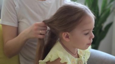 Otantik, genç, beyaz bir kadın annesi, çocuk örgüleriyle ilgileniyor sarı saçlı sarı saçlı kız örgüleri, gri kanepe üzerinde. Örgü örme işlemi. Moda ve güzellik, annelik kavramı.