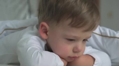 Beyaz bir yatakta rahat bir şekilde uyanan beyaz tenli, küçük, şirin, beyaz bir anaokulu çocuğunun otantik yakın çekimleri çene üzerinde duruyor. Çocuk bakımı, Uyuyan Bebek, Çocukluk, Ebeveynlik, Yaşam Konsepti