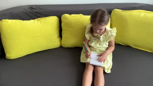 Portré Vicces kislány sárga ruhában mosolygós gyerek ír ceruzával jegyzetfüzetben ül a kanapén otthon. aranyos nő, csinos arccal a szürke kanapén. gyermekkor, távoktatási koncepció — Stock videók