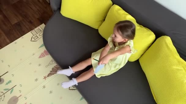 Sarı elbiseli, gülümseyen çocuk evdeki kanepede oturan kalemle yazıyor. Gri kanepede güzel yüzlü tatlı bir bayan çocuk. çocukluk, uzaklık öğrenme kavramı — Stok video