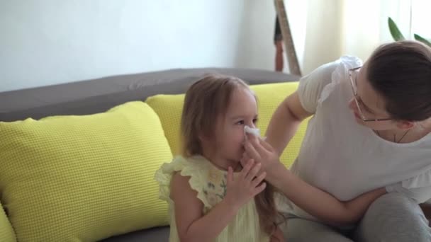 Średni plan autentyczne młody biały kobieta mama opieka nad dzieckiem pomaga wydmuchać nos w serwetce. Opieka macierzyńska chorej córki w żółtej sukience na szarej sofie. Medycyna i zdrowie, koncepcja koronawirusa — Wideo stockowe
