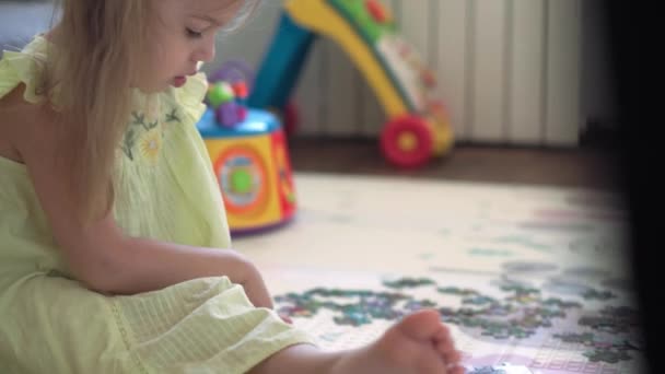 Autêntico bonito menina pré-escolar criança feliz coloca quebra-cabeça no chão de perto. criança pequena jogar sozinho jogos educativos para habilidades motoras perto de sofá cinza amarelo. Infância, família, conceito de isolamento. — Vídeo de Stock