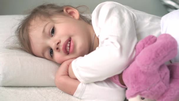 확실하게 귀여운 코카서스 꼬마 미취학 아동 소녀는 곰 곰 이 흰색 침대에서 카메라를 보고 있는 것을 보고 잠에서 깨어날 때 잠 이 들었다. 자녀 양육, 잠 자는 아기, 어린 시절, 부모로서의 삶의 개념 — 비디오