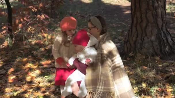 Aito söpö nuori nainen pikkulapsi vauva ja pieni lapsi tyttö kääritty huopa laskenut keltaiset lehdet syksyllä puistossa tai metsässä. Äiti huolehtii lapsista. Luonto, lapsuus, äitiyden käsite — kuvapankkivideo