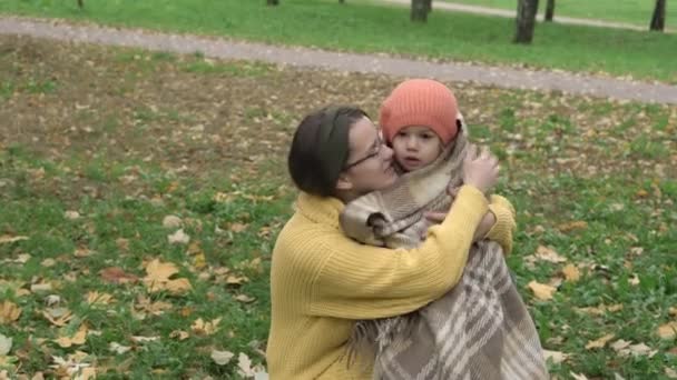 La infancia, la familia, la maternidad, el concepto de otoño - joven hermosa linda chica con el pelo largo y suelto oscuro en suéter amarillo envuelve en la niña a cuadros en boina naranja en el prado con hojas caídas en el parque — Vídeo de stock