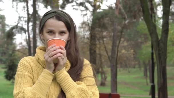Copilăria, familia, conceptul de toamnă - tânără fată drăguță frumoasă, cu păr lung și închis la culoare în pulover galben băuturi care țin ceașcă de hârtie portocalie cu ceai de garach sau cafea și zâmbește uitându-se la cameră — Videoclip de stoc