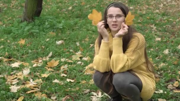 Kindheit, Familie, Herbstkonzept - junge hübsche Mädchen mit langen lockeren dunklen Haaren in orangefarbenem Pullover sitzen auf Gras und bedecken das Gesicht mit zwei gelben Ahornblättern und lächeln in die Kamera — Stockvideo