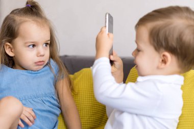 Mutlu ve üzgün küçük anaokulu kardeşleri küçük erkek ve kız kardeş sarı gri kanepede video konferansı hakkında konuşuyorlar. Oğlan, üzgün kızın resimlerini akıllı telefonla çekiyor. Çocukluk, teknoloji konsepti