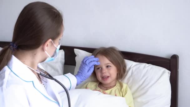 Αυθεντικό χαριτωμένο μικρό παιδί νηπιαγωγείο άρρωστο κορίτσι κοιτάζοντας κάμερα στο λευκό κρεβάτι. Καυκάσια νεαρή γυναίκα νοσοκόμα ή γιατρός παιδίατρος εξετάζει το μωρό στο νοσοκομείο. ιατρική και υγεία, έννοια covid-19 — Αρχείο Βίντεο