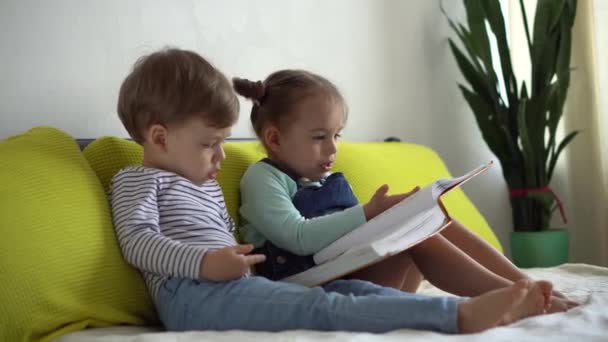 Persahabatan, keluarga, pendidikan Dua anak prasekolah tersenyum, balita membaca buku cerita dongeng yang menarik besar di tempat tidur kuning. Saudara kembar kecil pembaca bersenang-senang, anak-anak bahagia di karantina di rumah — Stok Video