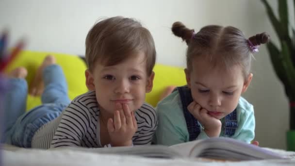 Amizade, família, educação Dois pré-escolares sorridentes, crianças pequenas ler grande livro interessante de contos de fadas na cama amarela. Irmãos pequenos gêmeos leitor se divertir, crianças felizes em quarentena em casa — Vídeo de Stock