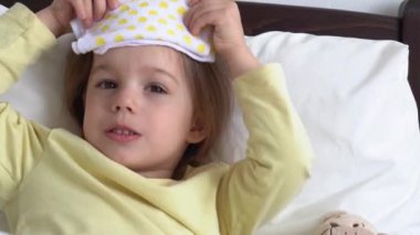 Otantik şirin küçük hasta anaokulu kızı alnına kompres uygular ve yatak odasındaki ya da hastane koğuşundaki beyaz yatağa gülümser. Tıp ve sağlık, çocukluk, covid-19 kavramı.