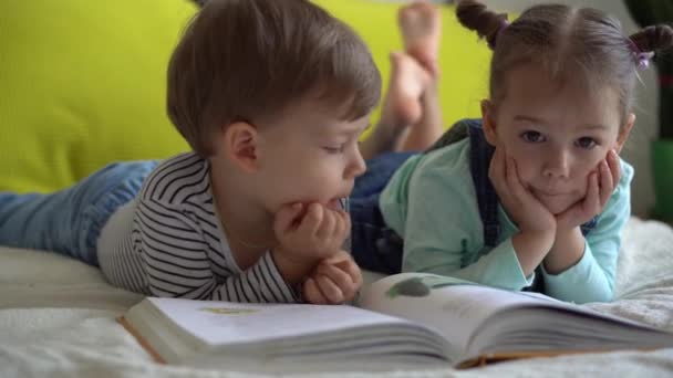Freundschaft, Familie, Bildung - zwei lächelnde Vorschulkinder lesen auf gelbem Bett ein großes, interessantes Märchenbuch. Geschwister kleine Zwillinge haben Spaß, glückliche Kinder zu Hause in Quarantäne — Stockvideo