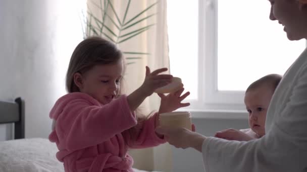 Autêntico bonito jovem mulher aplicando máscara facial de bebê menina. mãe com 2 filhos filhas diferentes idades em roupões de banho após o banho na cama branca janela oposite. Spa, maternidade, infância, conceito de família — Vídeo de Stock