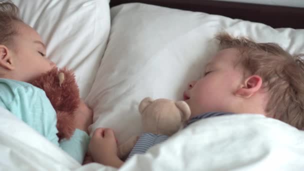 Authentisches Porträt niedlichen kaukasischen kleinen Vorschulgeschwistern Baby Junge und Mädchen im blauen Schlaf mit Teddybär auf weißem Bett. Mittags ruhen die Kinder. Pflege, Medizin und Gesundheit, Kindheit, Lebenskonzept — Stockvideo