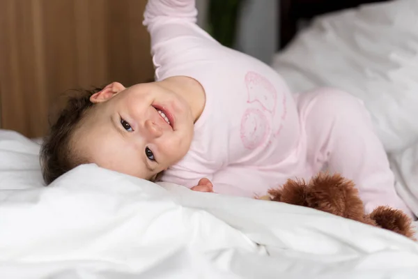 Подлинный портрет белый маленький пухленький младенец девочка или мальчик в розовом сонный, проснувшись глядя на камеру улыбаясь милые и гримасы в белой постели. Уход за детьми, Детство, Родительство, концепция жизни — стоковое фото