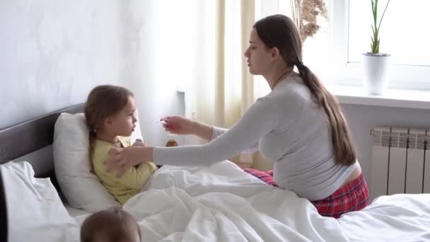 Jonge blanke vrouw verpleegkundige of arts onderzoekt baby meisje meet temperatuur met thermometer onder arm op wit bed. Mama zorgt voor ziek kind. geneeskunde en gezondheid, moederschap, covid-19-concept — Stockvideo