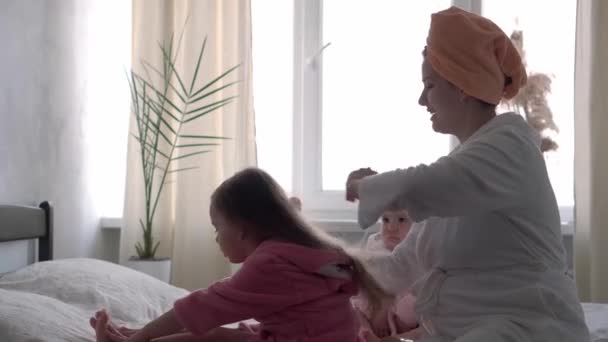 Auténtica joven linda peinando el pelo largo de la niña. mamá con 2 hijos hijas de diferentes edades en batas de baño después del baño en la ventana oposite cama blanca. Spa, maternidad, infancia, concepto familiar. — Vídeo de stock