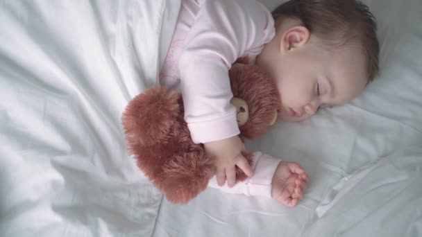 Authentisches Porträt niedlichen kaukasischen kleinen Säugling pummelig Baby Mädchen oder Junge im rosa Schlaf mit Teddybär auf weißem Bett. Mittags ruhen die Kinder. Pflege, Schlafkind, Kindheit, Elternschaft, Lebenskonzept — Stockvideo