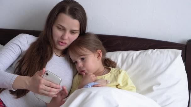 Kaukasische Familie kleine Kinder Mädchen und junge Frau Mutter sprechen in Videokonferenz per Smartphone zusammen zu Hause mit glücklichem Lächeln auf weißem Bett. Technologie, Lebensstil, Nutzungskonzept für Mobiltelefone — Stockvideo