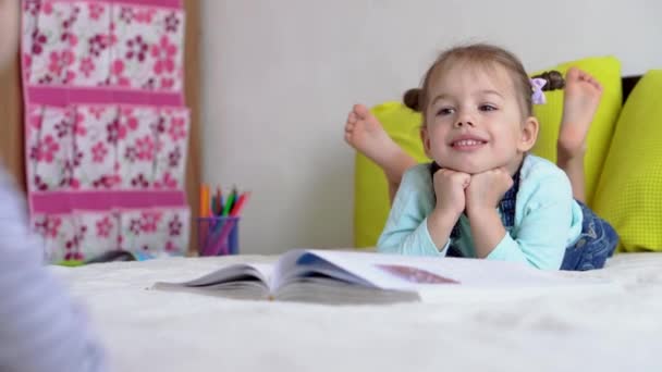 Дружба, сім'я, освіта - одна розумна усміхнена дошкільнята, маленька дівчинка читає велику цікаву книгу казок на жовтому ліжку. маленький милий читач розважається, щаслива дитина на карантині вдома — стокове відео