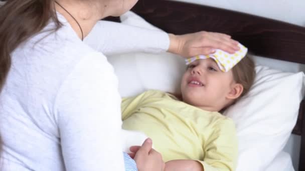 Nahaufnahme junge kaukasische Krankenschwester oder Arzt Kinderarzt untersucht Baby Mädchen legt Kompresse auf die Stirn auf weißem Bett. Mama kümmert sich um krankes Kind. Medizin und Gesundheit, Mutterschaft, Covid-19-Konzept — Stockvideo