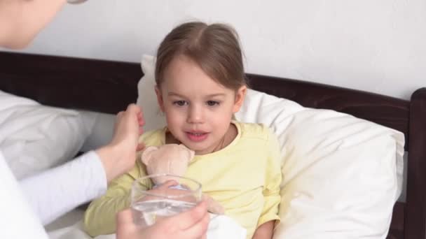 Κοντινό πλάνο νεαρή λευκή γυναίκα νοσοκόμα ή γιατρός παιδίατρος εξετάζει κοριτσάκι δώσει ποτό ποτήρι νερό στο λευκό κρεβάτι. Η μαμά φροντίζει το άρρωστο παιδί. ιατρική και υγεία, μητρότητα, covid-19 έννοια — Αρχείο Βίντεο