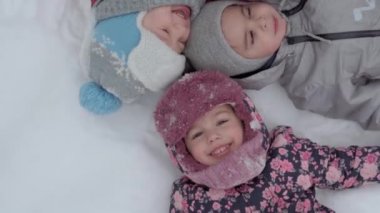 Tatil, aile kavramı - yavaş çekim otantik 3 mutlu anaokulu çocuğu erkek ve kız kardeşler başlarını birbirine yaslayarak sırt üstü uzanıyorlar. Soğuk mevsimde kar yağışı, kış parkında açık havada.
