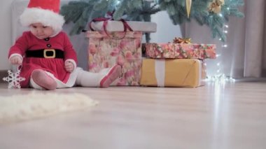 Noel Baba şapkası takan, kırmızı elbiseli, mutlu, mutlu, tombul bir kız çocuğu. Yeni yıl atmosferini kutlarken evdeki Noel ağacının yanında iyi eğlenceler. Çocukluk, bayram, kış konsepti