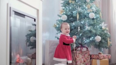 Noel Baba şapkası takan, kırmızı elbiseli, mutlu, mutlu, tombul bir kız çocuğu. Yeni yıl atmosferini kutlarken evdeki Noel ağacının yanında iyi eğlenceler. Çocukluk, bayram, kış konsepti