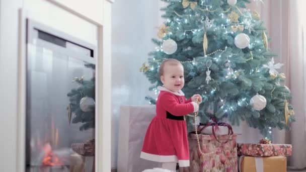 Auténtico lindo feliz alegría regordete niña con sombrero de santa y vestido rojo sonrisa jugar divertirse celebrando ambiente festivo año nuevo cerca del árbol de Navidad en casa. Infancia, día de fiesta, concepto de invierno — Vídeos de Stock