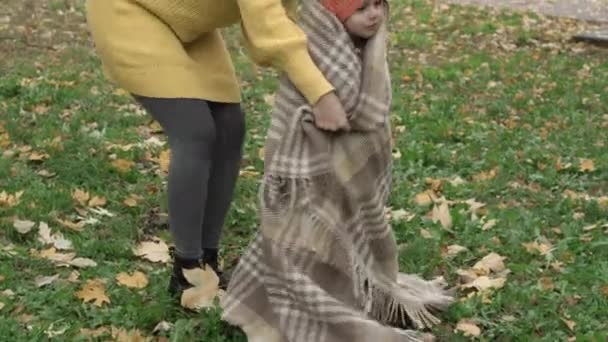 Childhood, family, motherhood, autumn concept - young beautiful cute girl with long loose dark hair in yellow sweater wraps in plaid little girl in orange beret on meadow with fallen leaves in park — Stock Video