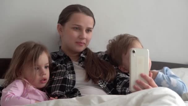 Authentieke jonge vrouw moeder van veel kinderen met baby jongen en meisje praten in video conferentie met smartphone samen thuis met glimlach op wit bed. technologie, levensstijl, concept van mobiel telefoongebruik — Stockvideo