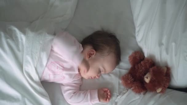 Authentisches Porträt niedlichen kaukasischen kleinen Säugling pummelig Baby Mädchen oder Junge im rosa Schlaf mit Teddybär auf weißem Bett. Mittags ruhen die Kinder. Pflege, Schlafkind, Kindheit, Elternschaft, Lebenskonzept — Stockvideo