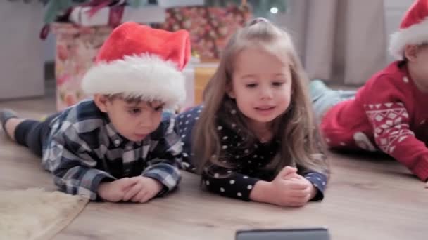 Autênticas crianças felizes Joy assistir desenhos animados no smartphone se alegrar pelo jogo árvore de Natal se divertir juntos. As crianças pré-escolares celebram a atmosfera festiva do Ano Novo em casa. Inverno, conceito de férias — Vídeo de Stock