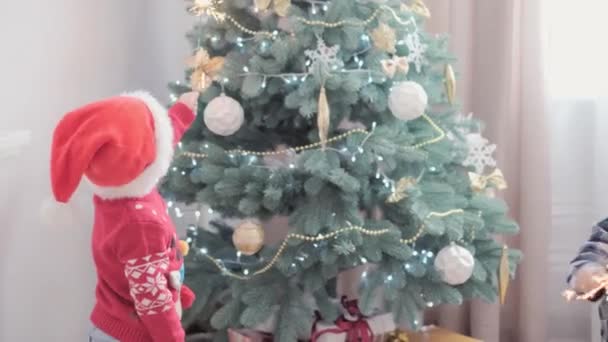 Auténtica alegría feliz niños con bengalas decoradas por el árbol de Navidad jugando divertirse juntos. El bebé preescolar celebra el ambiente festivo de Año Nuevo en casa. Invierno, vacaciones, concepto familiar — Vídeos de Stock