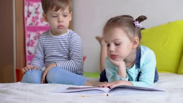 Amitié, famille, éducation - Deux enfants souriants d'âge préscolaire lisent un grand livre intéressant de contes de fées sur un lit jaune. Frères et sœurs petits jumeaux lecteur avoir du plaisir, enfants heureux en quarantaine à la maison — Video