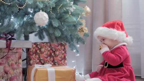 Аутентичная милая счастливая пухленькая девочка Джой в шапке Санта-Клауса и красной улыбке веселитесь празднуя новогоднюю праздничную атмосферу возле рождественской елки дома. Детство, праздник, зимняя концепция — стоковое видео