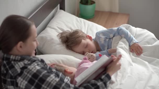 Otantik bir genç kadın ya da bebek bakıcısı yatmadan önce kitaptan çocuklara masal okur. Uyuyan çocuklara annem bakıyor. Beyaz yatakta uyuyan iki bebek. Anneler günü, annelik, çocukluk. — Stok video