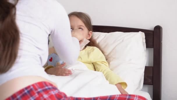 La joven enfermera caucásica o pediatra médica examina a una niña que le ayuda a sonarse la nariz en una servilleta en una cama blanca. Mamá cuida a un niño enfermo. medicina y salud, maternidad, concepto covid-19 — Vídeo de stock