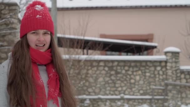 Slo-mo auténtica feliz linda mujer joven en sombrero rojo y bufanda mirando a la cámara en el patio cubierto de nieve en el día de Navidad. nevadas en temporada fría en el parque de invierno al aire libre. vacaciones, concepto de maternidad — Vídeos de Stock