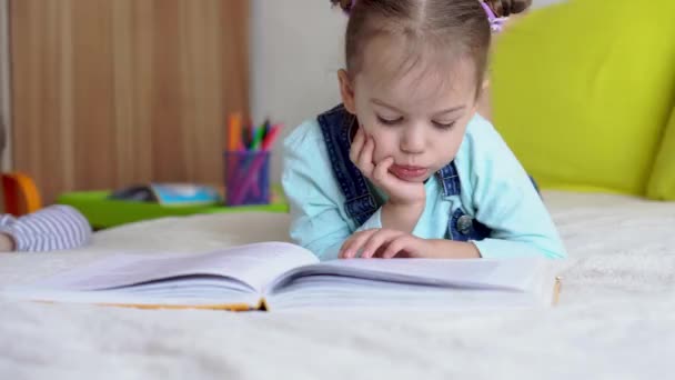 Дружба, сім'я, освіта - одна розумна усміхнена дошкільнята, маленька дівчинка читає велику цікаву книгу казок на жовтому ліжку. маленький милий читач розважається, щаслива дитина на карантині вдома — стокове відео