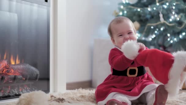 Auténtica niña gordita linda bebé en sombrero de santa y vestido rojo cerca de la chimenea con la madre joven celebrando feliz año nuevo cerca del árbol de Navidad. Infancia, paternidad, día de fiesta, concepto de invierno. — Vídeos de Stock