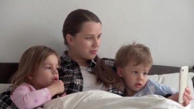 Beyaz yatakta gülümseyerek akıllı telefonla video konferansında konuşan çocuklu birçok çocuğun gerçek annesi genç bir kadın. teknoloji, yaşam tarzı, cep telefonu kullanımı kavramı