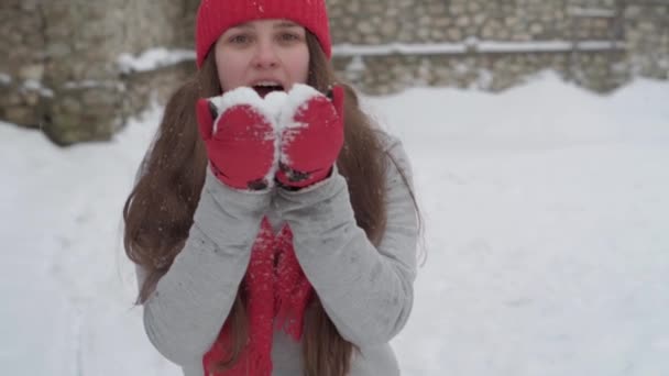 Slo-mo autentyczne słodkie szczęśliwa młoda kobieta z długimi pływające włosy w czerwonym kapeluszu i szalik patrząc na aparat z ładnym uśmiechem wziąć ciosy śniegu na płatki śniegu wymiotuje. macierzyństwo, koncepcja Dnia Matki — Wideo stockowe