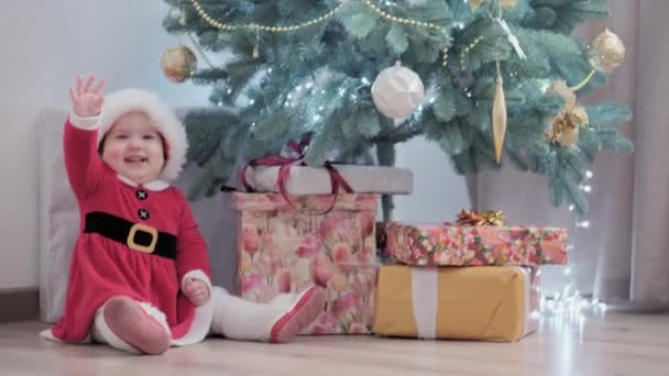 Аутентичная милая счастливая пухленькая девочка Джой в шапке Санта-Клауса и красной улыбке веселитесь празднуя новогоднюю праздничную атмосферу возле рождественской елки дома. Детство, праздник, зимняя концепция — стоковое видео