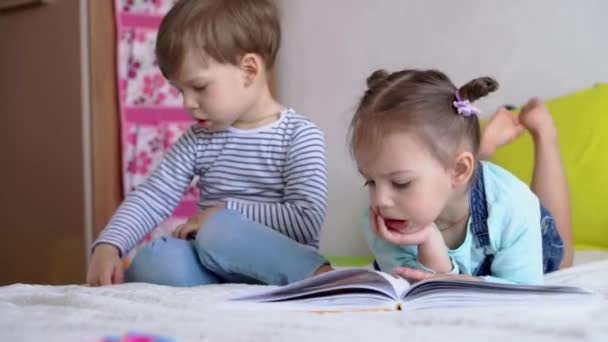友情、家族、教育- 2つの笑顔の幼稚園、幼児の子供たちは黄色のベッドの上でおとぎ話の大きな興味深い本を読んでいます。兄弟小さな双子のリーダーは家で隔離された楽しい幸せな子供を持っています — ストック動画